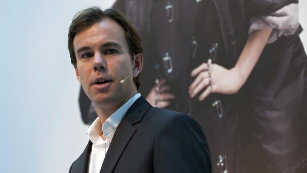 Ein Mann mit Anzug und Mikrofon spricht vor einem unscharfen Hintergrund.