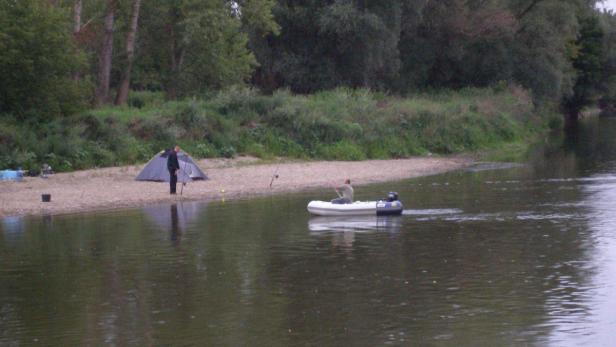 Ein Mann angelt vom Ufer aus, während ein anderer in einem Schlauchboot auf dem Fluss fährt.