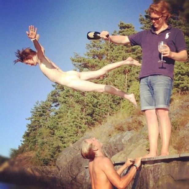 Eine Person springt nackt von einer Klippe, während andere mit Champagner und Glas zusehen.