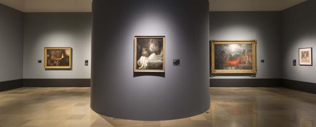 In einem Ausstellungsraum hängen mehrere Gemälde an den grauen Wänden.