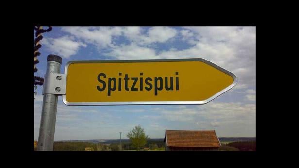 Ein gelbes Ortsschild mit der Aufschrift „Spitzispui“ vor einem bewölkten Himmel.
