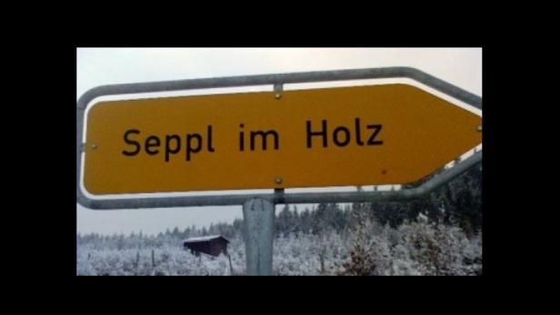 Ein gelbes Schild mit der Aufschrift „Seppl im Holz“ vor einer verschneiten Landschaft.
