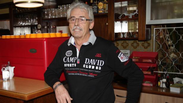 Ein Mann mit Brille und Schnurrbart lehnt an einer roten Kaffeemaschine in einem Café.