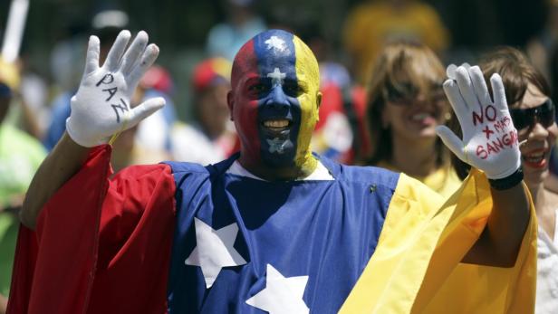 Ein Mann mit venezolanischer Flagge und Gesichtsbemalung demonstriert mit beschrifteten Handschuhen.