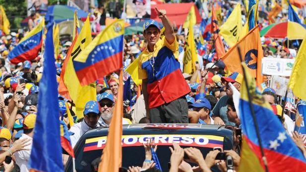 Ein Mann in den Farben Venezuelas steht auf einem Auto, umgeben von jubelnden Menschen und Flaggen.