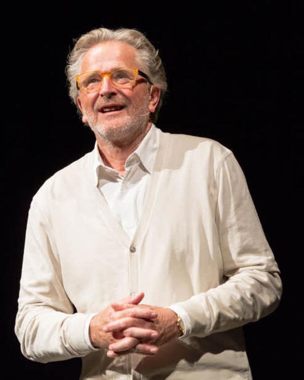 Ein älterer Mann mit Brille und hellem Cardigan steht vor einem dunklen Hintergrund.