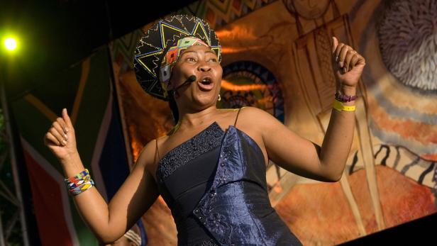 Eine Frau mit traditioneller Kopfbedeckung singt mit erhobenen Händen auf einer Bühne.