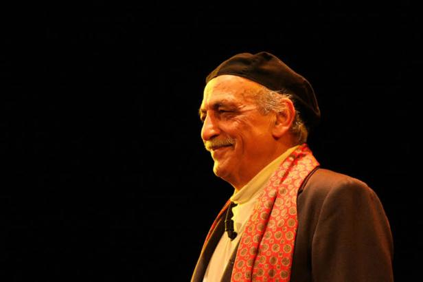 Ein lächelnder Mann mit Schnurrbart, Barett und gemustertem Schal vor schwarzem Hintergrund.