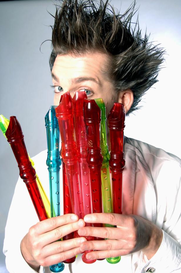 Ein Mann mit auffälliger Frisur hält mehrere bunte Blockflöten vor sein Gesicht.