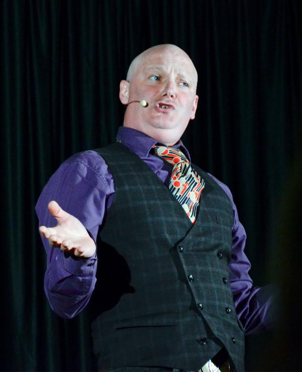 Ein Mann mit Mikrofon, lila Hemd, Weste und Krawatte gestikuliert vor einem schwarzen Vorhang.