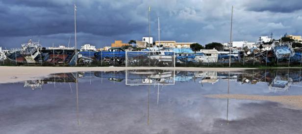 Ein Fußballtor steht vor einer Reihe alter, blauer Fischerboote, die sich in einer Wasserpfütze spiegeln.