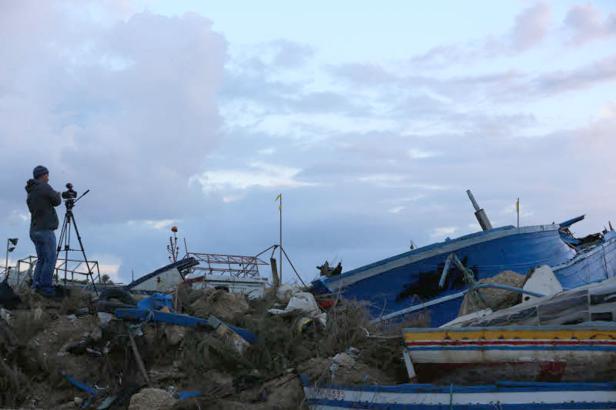 Ein Fotograf steht auf einem Trümmerhaufen und fotografiert ein zerstörtes blaues Boot.