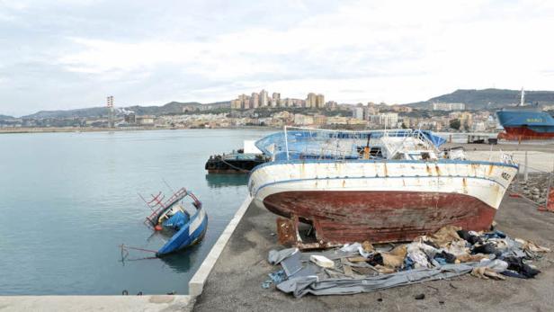 Ein beschädigtes Boot liegt teilweise im Wasser, ein weiteres steht an Land, im Hintergrund eine Stadt.