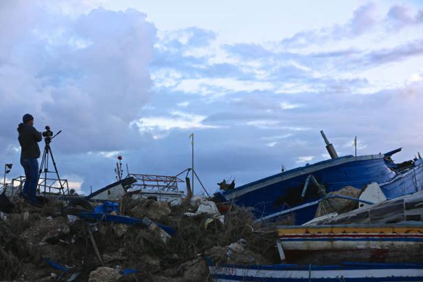 Ein Mann filmt mit einem Stativ die Überreste zerstörter Boote am Ufer.