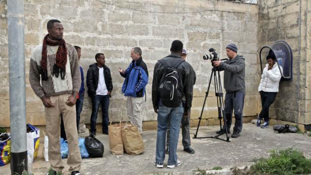 Eine Gruppe von Menschen steht vor einer Mauer, während ein Mann eine Kamera auf einem Stativ bedient.