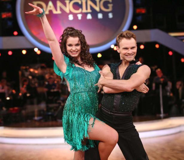 Ein Tanzpaar in glitzernden Kostümen tanzt vor dem „Dancing Stars“-Logo.