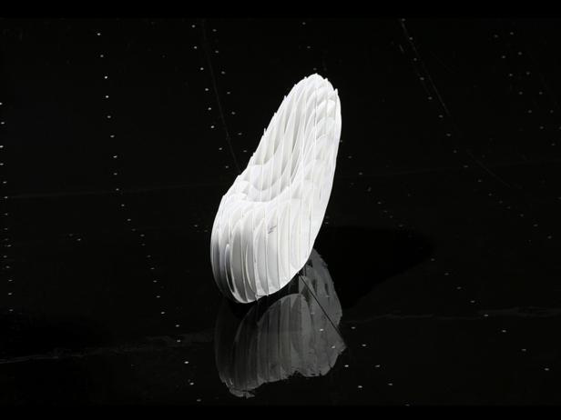 Ein abstrakter, weißer Schuh aus Papier steht auf einer glänzenden, schwarzen Oberfläche.