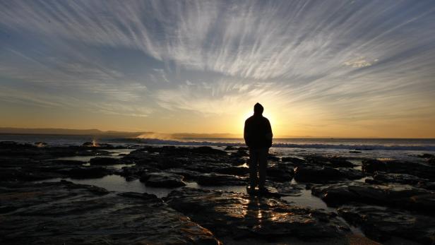 Eine Person steht bei Sonnenuntergang an einem felsigen Strand.