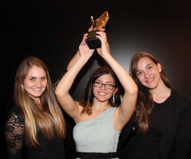 Drei junge Frauen posieren mit einer goldenen Trophäe vor einem dunklen Hintergrund.