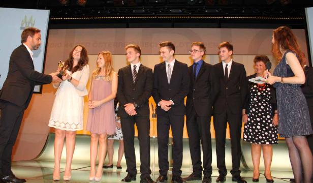 Eine junge Frau nimmt auf einer Bühne eine Auszeichnung entgegen, umgeben von anderen Personen.