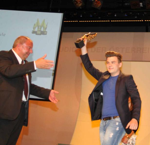 Ein junger Mann hält eine Trophäe in die Höhe, während ein anderer Mann ihm die Hand entgegenstreckt.