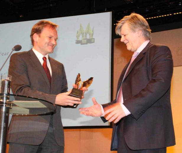 Ein Mann überreicht einem anderen eine goldene Trophäe auf einer Bühne.