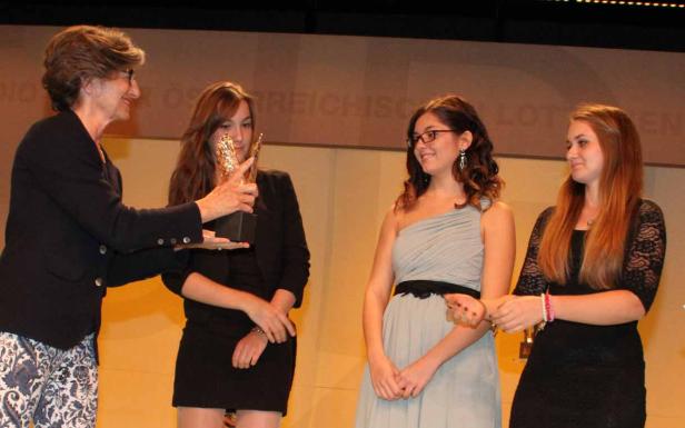 Eine Frau überreicht drei jungen Frauen eine Auszeichnung auf einer Bühne.