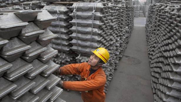 Ein Arbeiter mit Schutzhelm inspiziert gestapelte Aluminiumbarren in einer Fabrikhalle.