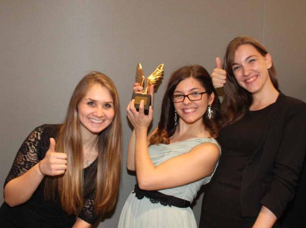 Drei Frauen posieren mit einem goldenen Preis und zeigen Daumen hoch.