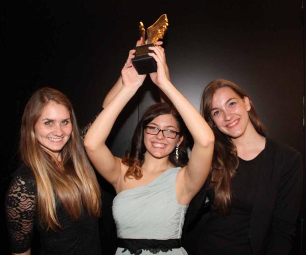 Drei junge Frauen halten gemeinsam eine goldene Trophäe in die Höhe.