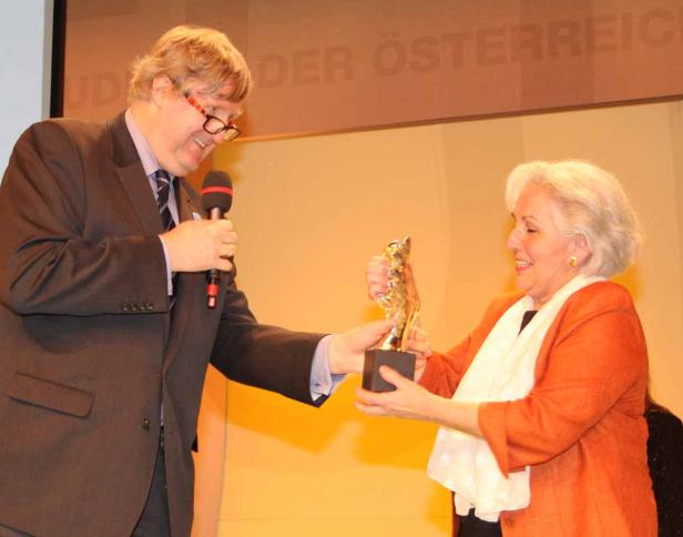 Eine Frau erhält eine goldene Statue von einem Mann mit Mikrofon.
