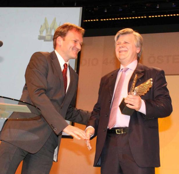 Zwei Männer in Anzügen auf einer Bühne, einer hält eine goldene Trophäe.