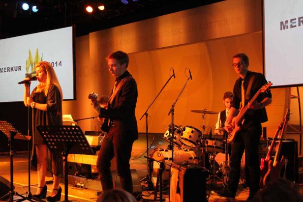 Eine Band spielt auf einer Bühne mit dem Schriftzug „Merkur 2014“ im Hintergrund.