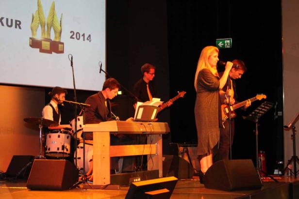 Eine Band spielt auf einer Bühne vor einer Leinwand mit der Aufschrift „2014“.