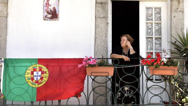 Eine ältere Frau steht auf einem Balkon mit portugiesischer Flagge und Blumenkästen.