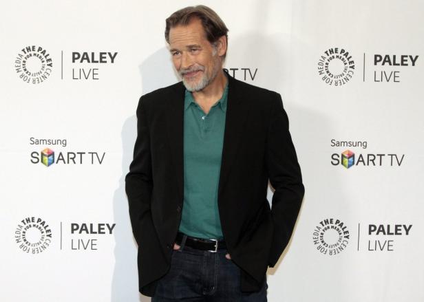 Ein Mann mit grauem Bart posiert vor einer Wand mit Logos.