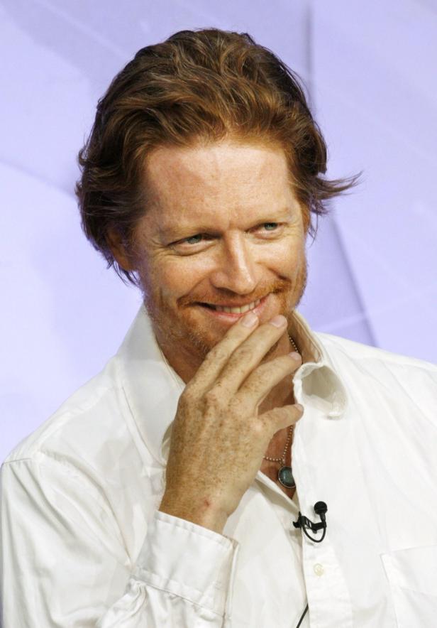 Ein lächelnder Mann mit roten Haaren und Sommersprossen berührt sein Kinn.