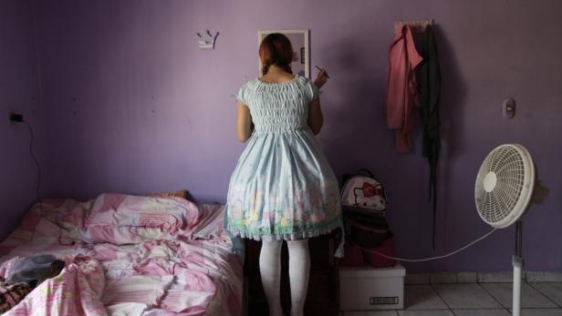 Eine junge Frau in einem hellblauen Kleid steht vor einem Spiegel und schminkt sich in ihrem Schlafzimmer.