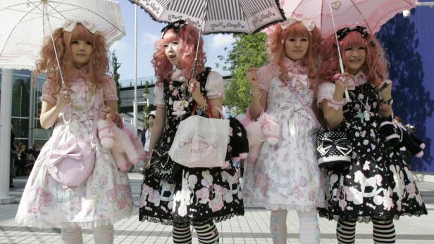 Vier junge Frauen im Lolita-Stil mit rosa Haaren und bunten Kleidern stehen unter Sonnenschirmen.