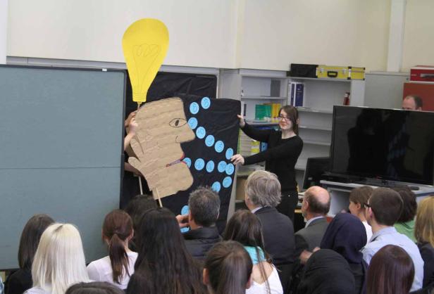 Eine Frau präsentiert ein Pappmodell eines Kopfes mit einer Glühbirne darüber vor einem Publikum.