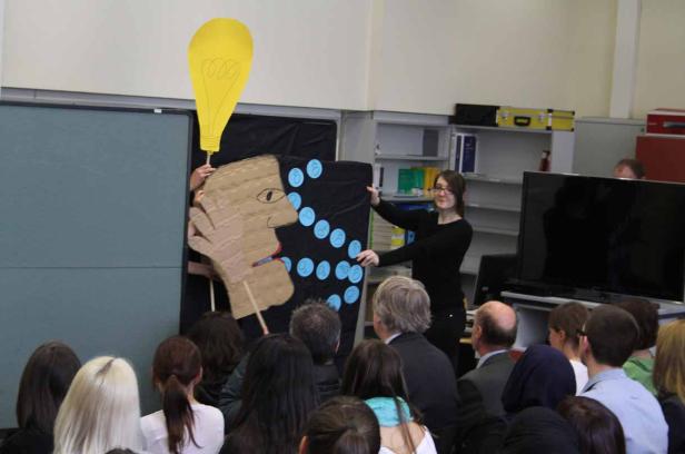 Eine Frau präsentiert vor Publikum eine Pappfigur mit Glühbirne.