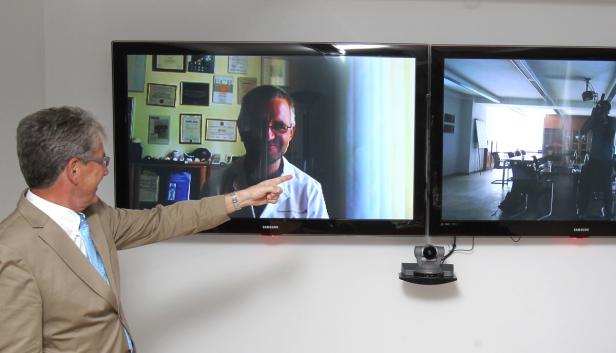 Ein Mann im Anzug zeigt auf einen Bildschirm mit einem Arzt während einer Videokonferenz.
