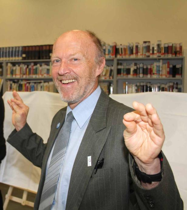 Ein lächelnder Mann im Anzug gestikuliert vor einem Bücherregal.