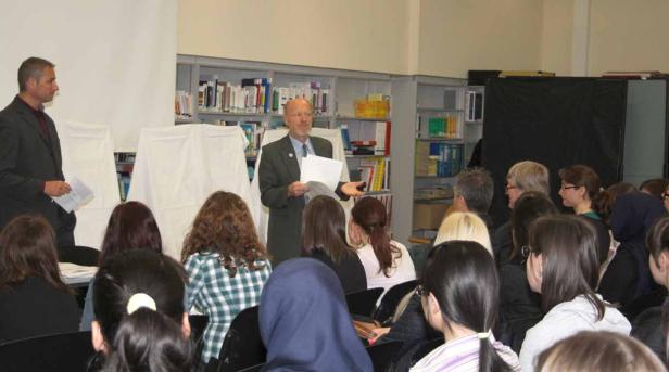 Zwei Männer sprechen vor einer Gruppe von Zuhörern in einem Raum mit Bücherregalen.