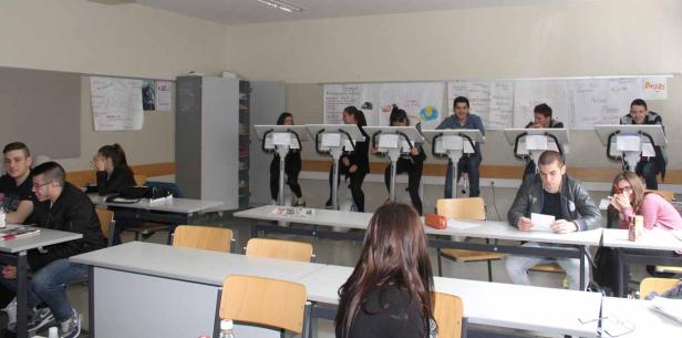 Schüler sitzen in einem Klassenzimmer, einige an Tischen, andere an Stehpulten mit Fahrrädern.