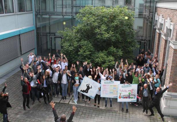 Eine Gruppe von Menschen springt mit erhobenen Händen vor einem Gebäude in die Luft.