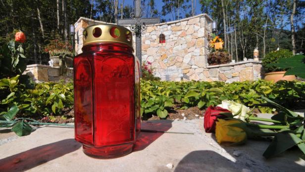 Eine rote Grabkerze und Rosen auf einem Grabstein vor einer Steinmauer mit Kreuz.