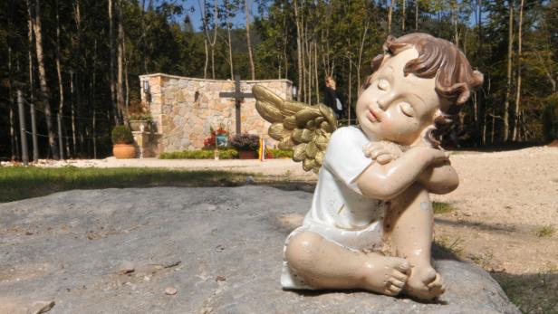 Eine Engelstatue sitzt auf einem Stein vor einer Gedenkstätte mit einem Kreuz.