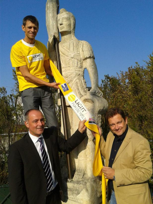 Drei Männer posieren mit einem gelben Banner der Organisation „Licht für die Welt“ vor einer Statue.