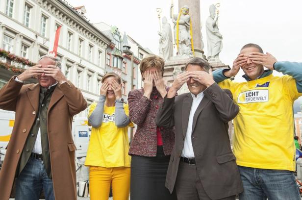 Fünf Personen verdecken sich auf einem Platz die Augen.